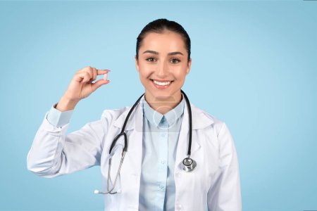 Photo for Friendly woman doctor in a white coat and stethoscope displays a small pill between her fingers, signifying medical treatment, blue background - Royalty Free Image
