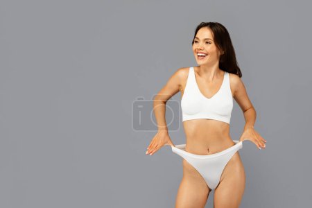 Photo for Laughing asian millennial lady in white underwear playfully pulling at underpants, looking to copy space, enjoy weight loss result, isolated on grey background. Joy, fit and body care - Royalty Free Image