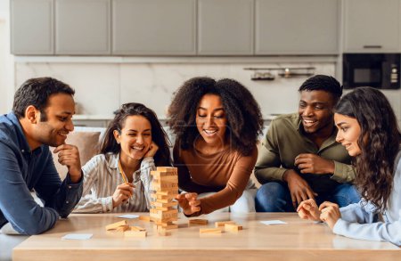 Foto de Jóvenes amigos multiétnicos jugando juego de mesa divertirse en casa cocina, la construcción de torre de madera y sonriendo. Estudiantes pasando tiempo juntos el fin de semana. Entretenimiento y juegos - Imagen libre de derechos
