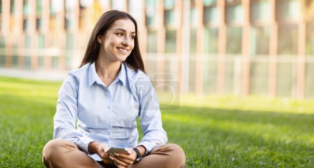 Photo for Happy arab young woman sits cross-legged on grass with smartphone, look at copy space, relaxed at spare time, chatting. Device, typing in social networks outdoors, panorama - Royalty Free Image