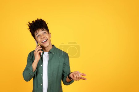 Foto de Joven afroamericano positivo hablando por teléfono celular, mirando espacio de copia para publicidad y gestos, aislado en el fondo amarillo del estudio. Concepto de comunicación - Imagen libre de derechos