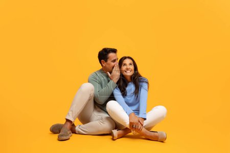 Photo for Seated man whispers into woman ear, smiling with look of shared secrecy and joy, capturing candid moment of connection against yellow backdrop. Gossip and joyful connection - Royalty Free Image