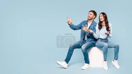 Téléchargez les photos : Homme et femme excités en denim pointant vers le côté assis sur un cube blanc, plein d'énergie et d'enthousiasme sur un fond bleu, espace libre - en image libre de droit