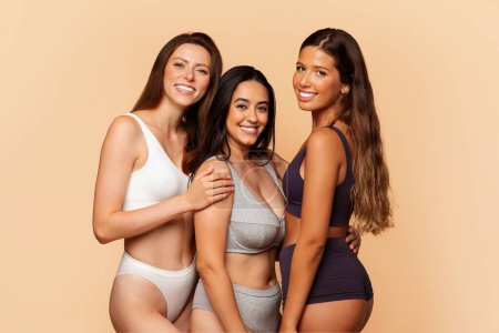 Photo for Three cheerful multiracial women in underwear embrace and smile against warm beige background, depicting camaraderie and diverse beauty. Friendship, diversity, joy and lifestyle - Royalty Free Image