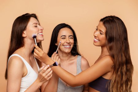 Photo for Smiling millennial international women in underwear apply nude makeup with brushes to friend, enjoy beauty care, isolated on beige background, studio. Cosmetics, fashion ad and offer - Royalty Free Image