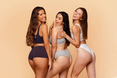 Téléchargez les photos : Trois jeunes femmes multiraciales dans des vêtements de sport élégants souriant et posant ludique, respirant la confiance et la camaraderie, s'amuse sur fond beige. Confiance, bien-être, amitié, mode de vie actif - en image libre de droit