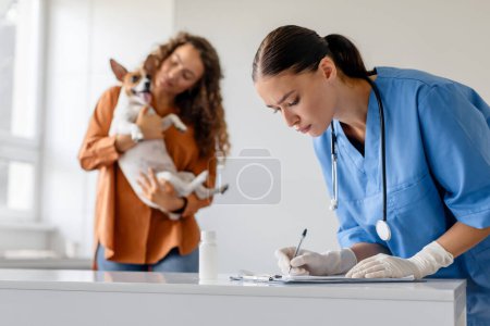 Photo for Focused veterinarian writes on clipboard while pet owner playfully holds her Jack Russell Terrier at veterinary check-up in a white room - Royalty Free Image