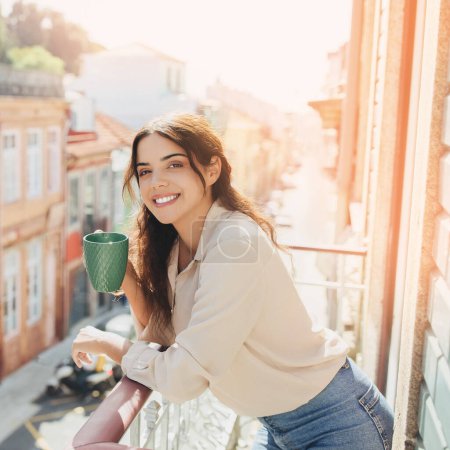 Téléchargez les photos : Joyeux jeune femme avec tasse de café debout sur un balcon ensoleillé, heureuse jolie femme européenne dégustant une boisson à la caféine, se détendre à la maison avec une tasse de thé, profiter de la vue et sourire à la caméra - en image libre de droit