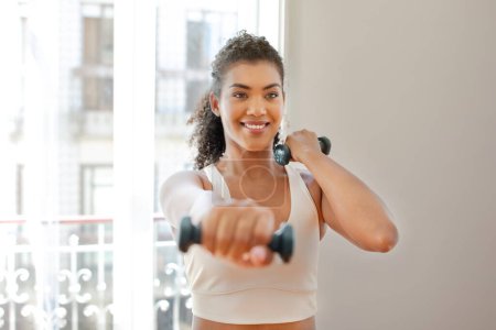 Photo for Active athletic young woman holds dumbbells with a smile, exercising in her living room. Motivated fitness lady in activewear demonstrates strength and balance, flexing arms muscles during workout - Royalty Free Image
