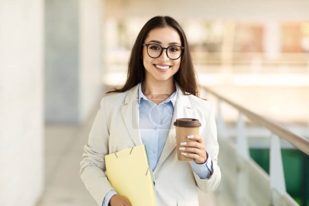Photo for Smiling young businesswoman with glasses, holding a coffee cup and yellow folders, ready for a productive workday. Preparedness and enthusiasm in a business setting - Royalty Free Image