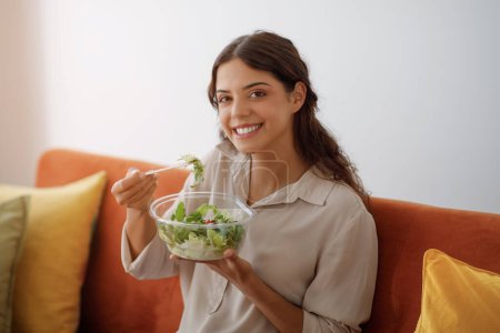 Téléchargez les photos : Jeune femme heureuse mangeant de la salade de légumes tout en étant assise sur le canapé à la maison, souriant belle femme ayant un déjeuner sain, appréciant les légumes frais, relaxant dans l'intérieur du salon, espace de copie - en image libre de droit