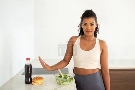 Téléchargez les photos : Pas de malbouffe. Motivé femme sportive mince geste arrêt à burger malsain et boisson sucrée douce dans la cuisine, le choix de salade de légumes frais pour le déjeuner. Choix d'aliments sains, régime de perte de poids - en image libre de droit