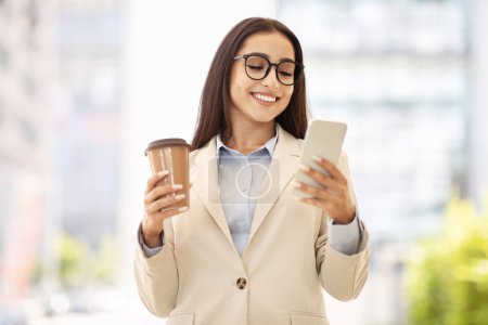 Foto de La empresaria contenta con una elegante chaqueta beige sostiene una taza de café y un teléfono inteligente, que se dedica a leer la pantalla al aire libre. Multitarea moderna y uso de tecnología sobre la marcha - Imagen libre de derechos