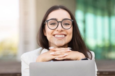 Foto de Divertida alegre mujer árabe joven con gafas descansa su barbilla en las manos por encima de la computadora portátil, exudando positividad y satisfacción laboral, se divierte. Disfrutar del trabajo y el ambiente de trabajo positivo, de cerca - Imagen libre de derechos