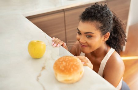 Foto de Elección de comida. Señora en forma hambrienta mirando hamburguesa malsana y manzana sana, eligiendo su comida cerca de la mesa de la cocina en el interior. Mujer deportiva elige entre una nutrición equilibrada y desequilibrada - Imagen libre de derechos