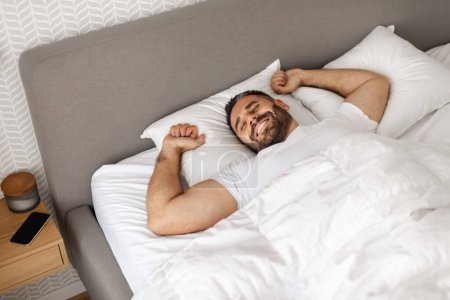 Téléchargez les photos : Bien reposé, l'homme d'âge moyen se réveille tranquillement en étirant les bras dans son lit à la maison, profitant du réveil matinal dans son intérieur moderne de chambre à coucher. Bon concept de sommeil et de loisirs - en image libre de droit