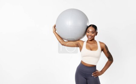 Téléchargez les photos : Entraînement de fitness. Femme noire athlétique en tenue de sport tient la balle de stabilité, souriant à la caméra montrant sa force et son équilibre, posant sur fond de studio blanc. Espace vide pour le texte - en image libre de droit