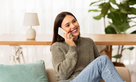 Photo for Positive young brunette lady enjoying phone conversation at home. Happy beautiful woman relaxing on couch in living room, talking on smartphone with lover, looking at copy space and smiling - Royalty Free Image