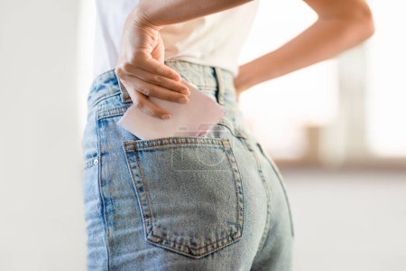 Photo for Closeup of lady putting menstrual pad in jeans pocket, caring for her comfort and protection during menstruation period, standing at home. Healthy organic female sanitary products - Royalty Free Image
