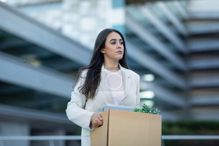Téléchargez les photos : Triste femme d'affaires hispanique portant une boîte en carton avec ses biens, quittant l'immeuble de bureaux après la mise à pied, reflétant les défis économiques du licenciement des entreprises et du chômage - en image libre de droit