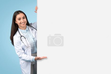 Foto de Alegre doctora en bata blanca muestra un espacio en blanco para publicidad médica o contenido informativo, presentando cartelera sobre fondo azul - Imagen libre de derechos