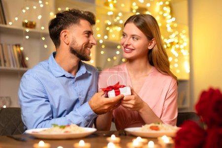 Photo for Man with cheerful smile presents small gift box to pleasantly surprised woman, enhancing the joy of their intimate candlelit dinner - Royalty Free Image