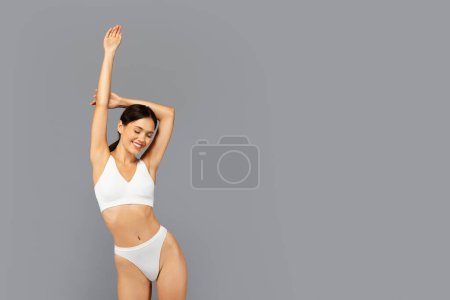 Téléchargez les photos : Joyeuse femme millénaire asiatique en sous-vêtements blancs souriant et fermant les yeux, profiter de temps libre, soins du corps, isolé sur fond de studio gris. Bonheur et bien-être physique - en image libre de droit