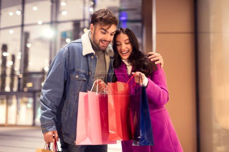 Photo for Young couple joyfully displays their Valentines Day shopping, peeking inside bags standing together outside a city mall at winter night. Festive sales and discounts concept - Royalty Free Image