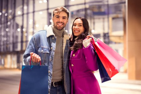 Photo for New Year Shopping Spree. European couple in winter wear smiles broadly holding colorful paper shopper bags outside of brightly lit urban mall, posing with their Christmas shopping purchases - Royalty Free Image