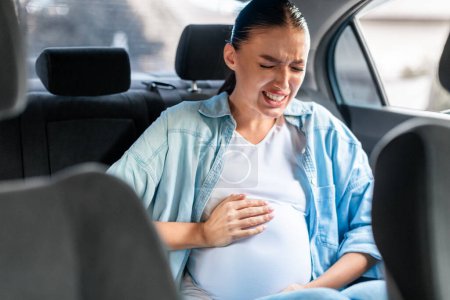 Foto de Mujer embarazada en el coche que sufre de dolores de parto agarrando su abdomen mientras está sentado en el asiento trasero de auto. Problemas de salud y transporte durante el embarazo. Enfoque selectivo - Imagen libre de derechos