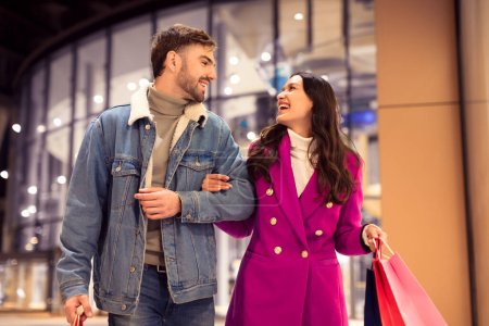 Téléchargez les photos : Jeune couple européen aime faire du shopping d'hiver festif, riant ensemble tout en marchant en se tenant la main avec des sacs de cadeaux de vacances, à l'extérieur du centre commercial urbain la nuit d'hiver - en image libre de droit