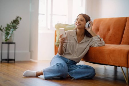 Photo for Favorite Playlist. Smiling Young Woman Wearing Wireless Headphones Listening Music On Smartphone While Relaxing At Home, Beautiful European Female Sitting On Floor In Living Room Interior, Copy Space - Royalty Free Image