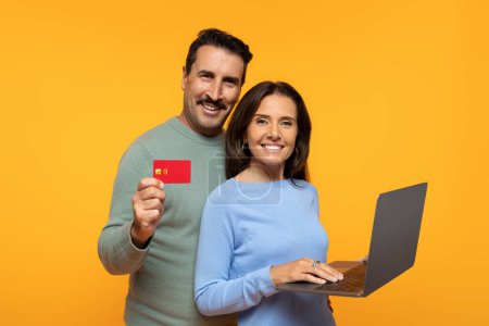 Foto de Pareja alegre se dedica a las compras en línea, el hombre que muestra la tarjeta de crédito, los tipos de mujer en el ordenador portátil, ambos sonriendo con confianza en un fondo naranja. Comercio electrónico y pagos digitales - Imagen libre de derechos