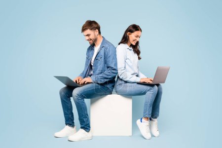 Photo for Concentrated young couple, dressed in stylish denim, working on laptops while sitting back-to-back on a white cube against a soft blue background - Royalty Free Image