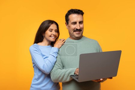 Foto de Hombre europeo sonriente en suéter y mujer miran juntos a la pantalla del portátil, señora descansando la mano sobre su hombro, ambos mostrando expresiones de interés y placer sobre fondo naranja - Imagen libre de derechos