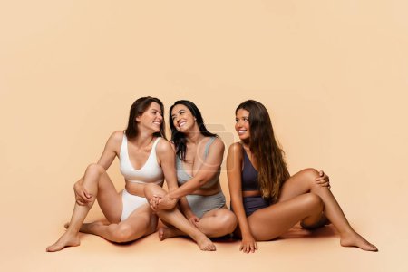 Téléchargez les photos : Trois femmes millénaires heureuses et diversifiées en sous-vêtements s'assoient sur le sol, apprécient la positivité du corps, les soins de beauté, s'amusent, isolées sur un fond de studio léger, pleine longueur. Amitié, confiance - en image libre de droit