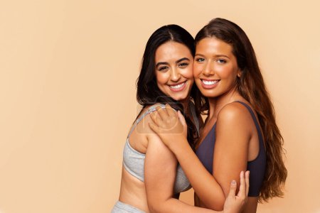 Photo for Two diverse positive young women in underwear, hugs, enjoy body care, isolated on beige studio background. Wellness, friendship, spa procedures, fit ad and offer, lifestyle - Royalty Free Image