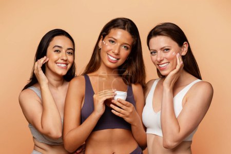 Téléchargez les photos : Trois jeunes femmes multiethniques en sous-vêtements montrent pot, mettre de la crème sur le visage, profiter de soins de beauté, l'hydratation à la maison sur fond de studio beige. Style de vie amusant, cosmétiques et spa, gros plan - en image libre de droit