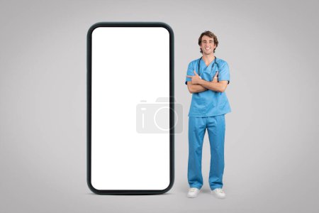 Photo for Cheerful man nurse, surgeon in blue uniform point finger at big phone with empty screen, isolated on gray background, full length. Healthcare services recommendation, ad and offer app - Royalty Free Image