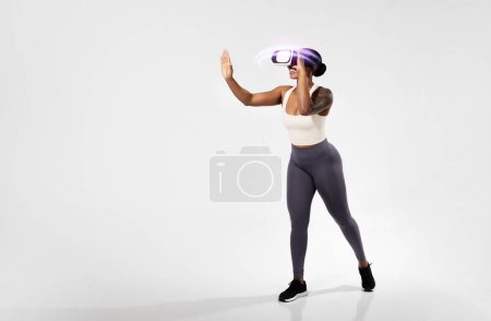 Téléchargez les photos : Jeune femme noire qui s'entraîne avec un casque de réalité virtuelle, explore le jeu de fitness VR, se tient debout sur fond de studio blanc. Plan complet, espace libre pour le texte - en image libre de droit