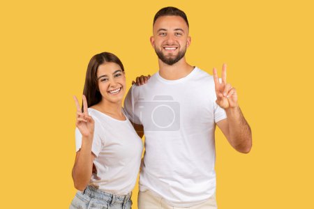 Photo for Radiant young european couple in white shirts showing peace signs with bright smiles, projecting a vibe of harmony and positivity gesture against a yellow studio backdrop - Royalty Free Image