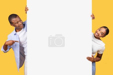 Photo for Enthusiastic black man and woman smiling and pointing to large blank banner they are holding, perfect for advertising or messages, against yellow backdrop - Royalty Free Image