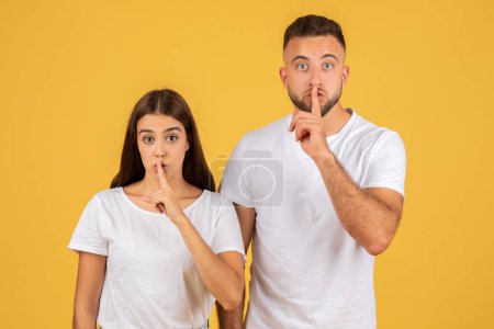 Téléchargez les photos : Surprised young european couple in white t-shirts with fingers on lips making a "shush" gesture, implying a need for silence or secrecy, against a vibrant yellow background - en image libre de droit