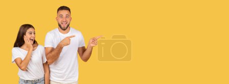 Foto de Excited happy young caucasian man and woman in white t-shirts point to the side with wide smiles, sharing a fun moment on a solid yellow studio background, panorama. Attention, good news, sale - Imagen libre de derechos