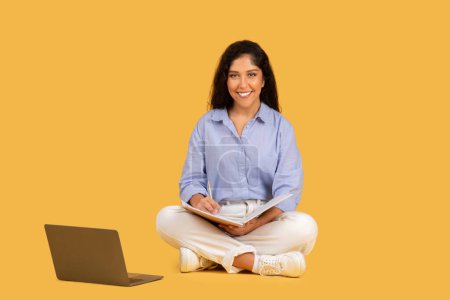 Téléchargez les photos : Focused young woman in casual attire sits with a notebook on her lap, beside an open laptop on a yellow background, symbolizing multitasking and productivity. Work, study, education - en image libre de droit