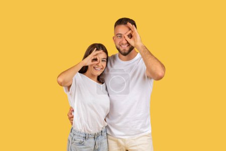 Photo for Playful young european couple in white t-shirts making fun gestures with their hands over one eye, standing close and smiling against a yellow studio background. Spare time, fun together - Royalty Free Image