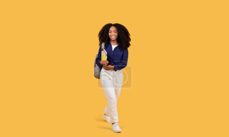 Photo for Cheerful black young lady with curly hair, exuding happiness, walks confidently with backpack and copybooks, on yellow background, depicting youth and education - Royalty Free Image