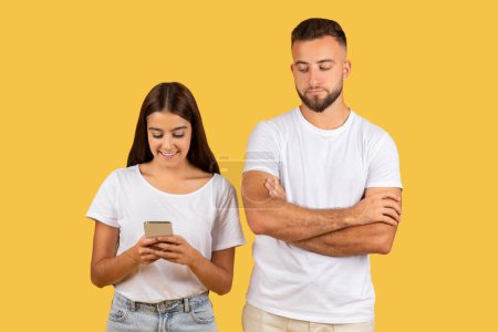 Téléchargez les photos : Smiling young woman in a white t-shirt engaged with her smartphone while a man in a white t-shirt stands beside her with arms crossed, looking away thoughtfully on a yellow background - en image libre de droit