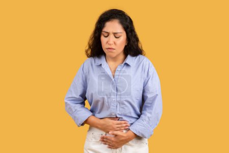 Téléchargez les photos : A young woman in a light blue shirt appears uncomfortable or in pain, holding her stomach with a distressed expression against a yellow background. Periods, health problems - en image libre de droit