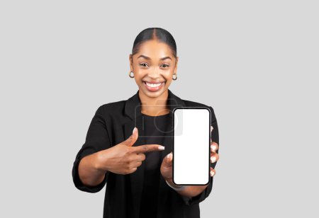 Foto de Smiling young african american woman in formal suit point finger at smartphone with empty screen, isolated on gray studio background. Business, work app, website recommendation - Imagen libre de derechos